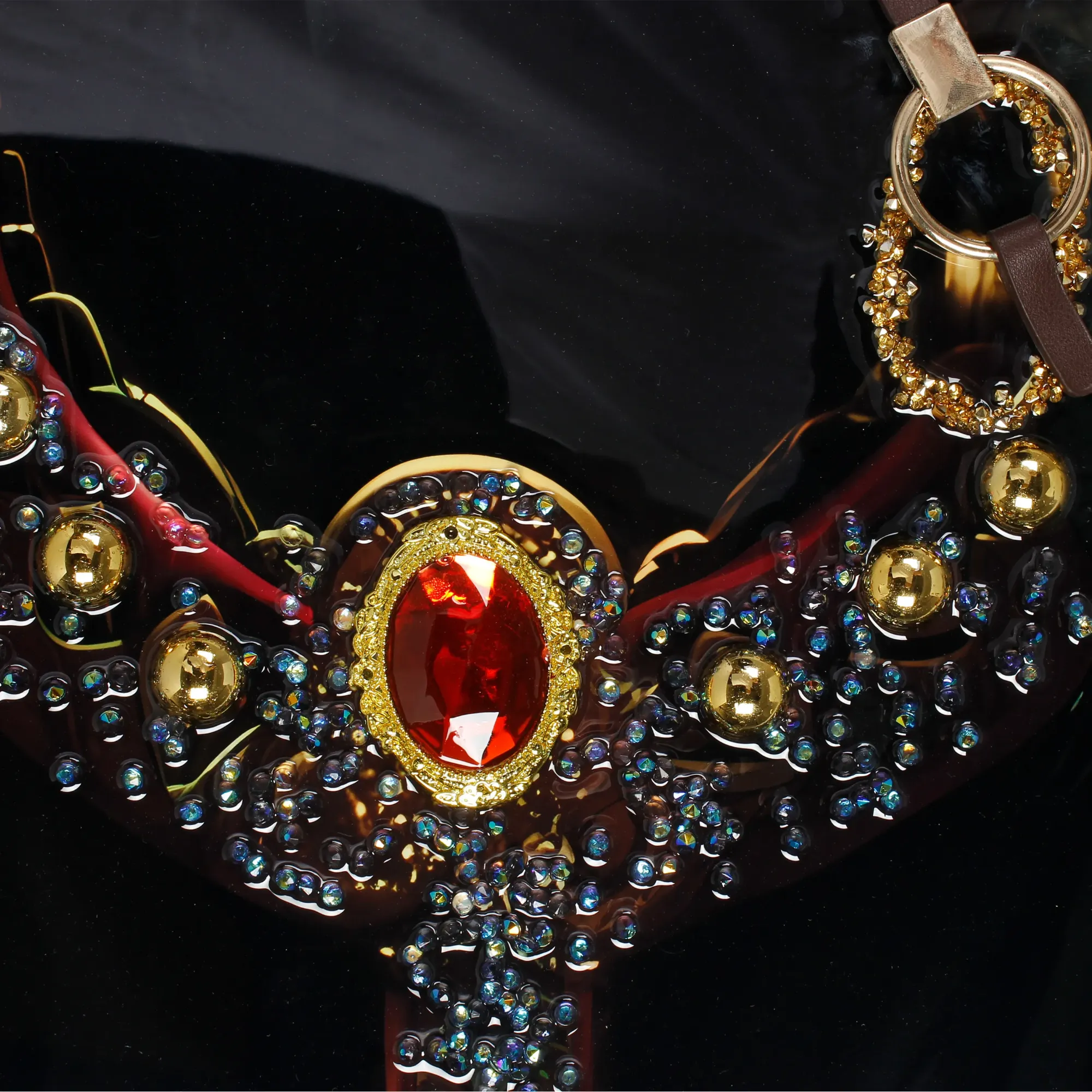  Extreme close-up of the black horse's ornate bridle featuring a large oval red gemstone set in a gold filigree frame, surrounded by small blue and clear crystal rhinestones, gold spherical studs, and dark red leather straps on a black background.